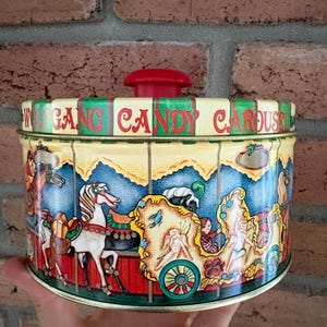 May include: A round metal tin with a red lid knob, featuring a colourful carousel illustration and the text "WOLFGANG CANDY CAROUSEL" printed in red on the lid rim. The design includes white horses, golden chariots, and figures.