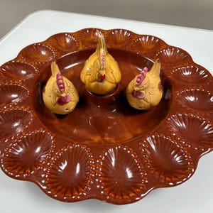 May include: A brown ceramic egg serving platter with a scalloped edge, holding three decorative chicken figurines. The chickens are yellow with red accents. The platter has individual egg compartments.