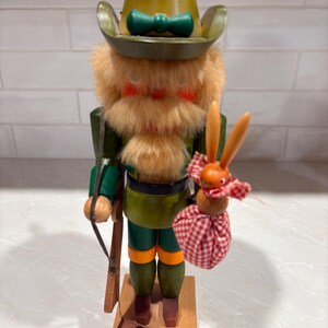May include: A wooden nutcracker figure wearing a green hat and outfit with a thick, light brown faux fur beard. The figure holds a small wooden rabbit toy attached to a red and white checkered fabric bundle. A brown tag hangs from the base.