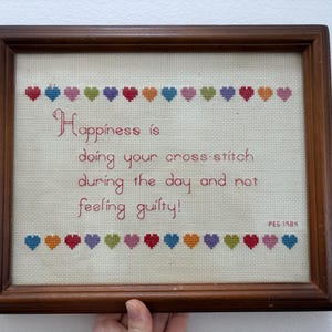 May include: Framed cross-stitch art with the quote "Happiness is doing your cross-stitch during the day and not feeling guilty!" The text is in red, with colorful heart accents. The frame is brown.