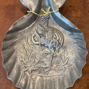 May include: A silver-toned metal tray shaped like a large scallop shell. The center features a raised relief design of a crane standing among aquatic plants. A thin yellow string is tied across the top section of the shell-shaped metal dish.