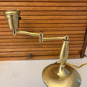 May include: A brass-toned desk lamp with an adjustable arm and a round base. The lamp has a classic design with a bulb socket at the end of the arm. The lamp is on a white surface with a wooden shutter background.
