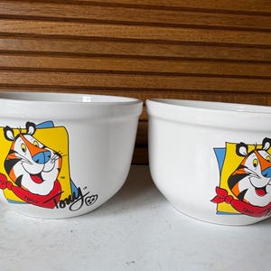 May include: Two white ceramic bowls with a cartoon tiger graphic. The tiger has orange and black stripes, a blue nose, and a red bandana. The bowls have the text "Tony" and the TM symbol.