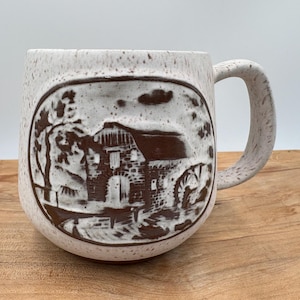 May include: A speckled ceramic mug with a brown relief design of a rural scene, including a building, trees, and a water wheel. The mug has a large handle and a rounded shape. The background is a light color.