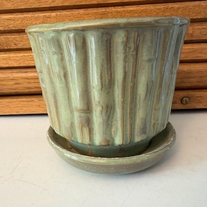 May include: A light green ceramic plant pot with a matching saucer. The pot features vertical ridges and a slightly flared top. The glaze has a mottled appearance with brown undertones, giving it a rustic look. Suitable for small plants.