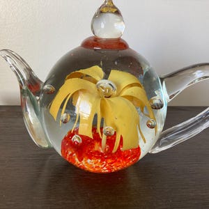 May include: A clear glass teapot with a decorative interior. The teapot features a yellow floral design and a red base. The handle and spout are also clear glass. The teapot is approximately 6 inches long.