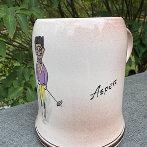May include: Cream-colored ceramic mug with a brown rim and handle. The mug features a skier illustration in purple and yellow, with the word "Aspen" in black script. The mug is a vintage style.