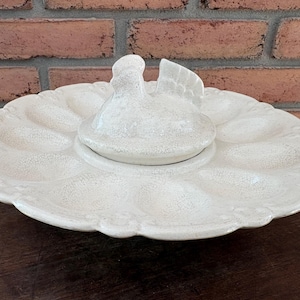 May include: A cream-colored ceramic egg serving platter featuring a central raised lid shaped like a hen. The circular tray has twelve molded indentations for eggs, a scalloped edge, and a textured, crackled glaze finish across the entire surface.