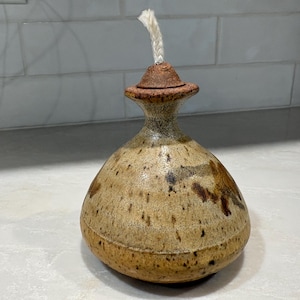 May include: A ceramic oil lamp with a rounded, bulbous base and a narrow neck. The exterior features a speckled tan and brown glaze with darker brown patches. A small, conical lid sits atop the neck, with a white cotton wick protruding from the centre.