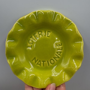 May include: A chartreuse ceramic ashtray with a ruffled edge. The center of the ashtray has the words "LOTERIE NATIONALE" embossed in a circular pattern. The ashtray is round and has a glossy finish.