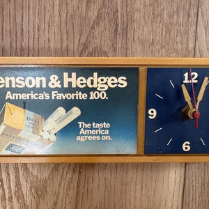 May include: A rectangular wall clock with a light brown frame. The left side features a Benson & Hedges advertisement with a cigarette pack graphic and text. The right side has a dark blue clock face with white numbers 3, 6, 9, 12 and gold hands.