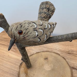 May include: A hand-carved wooden bird sculpture perched on a branch, with a wooden base. The bird has a detailed, textured surface with brown and white accents, a dark beak, and a round, orange eye. The sculpture is set against a light background.