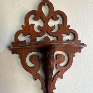 May include: A dark brown, ornate wooden wall shelf with a decorative, openwork design. The shelf has a small, curved surface for displaying items and is mounted on a wall. The shelf is approximately 10 inches wide.