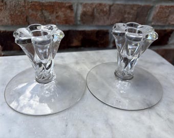 May include: Two clear glass candle holders with a flower-shaped top and a twisted stem design. The candle holders have a round base and are displayed on a marble surface. The background features a brick wall.