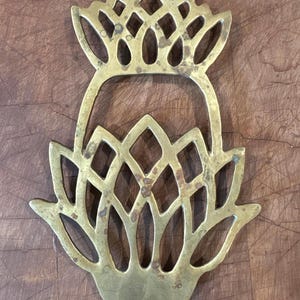 May include: A gold-toned brass trivet shaped like a pineapple with an openwork lattice design. The metal surface features a weathered patina with visible dark spots and a smooth, flat finish throughout the entire decorative kitchen accessory.