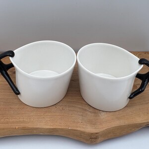May include: Two white ceramic mugs with black figure handles. The mugs are on a wooden serving tray. The handles are in the shape of a person. The mugs have a unique and modern design.