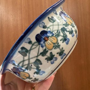 May include: A ceramic bowl with a cream-colored base featuring a hand-painted pattern of blue and yellow pansies connected by green vines. The bowl has a wide, rounded shape with a thick, solid dark blue rim around the top edge.