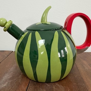May include: A ceramic teapot with a watermelon design. The body is dark green with light green stripes. It has a red handle, a green spout, and a green lid with a small green knob. The teapot is sitting on a wooden surface.