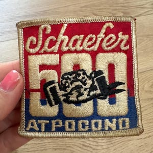 May include: A square embroidered patch featuring a red top section with the text Schaefer in gold script, a large gold 500 with a black race car graphic in the centre, and a blue bottom section with the text AT POCONO in gold block letters.