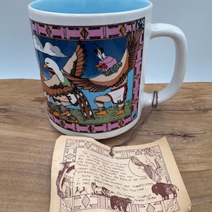 May include: White ceramic mug with a light blue interior and a handle. The mug features a colorful illustration of two birds in Native American attire, set against a blue sky and pink geometric border. A tag with text hangs from the handle.