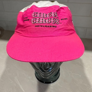May include: A bright pink baseball cap with a white top panel. The front features the text Circus Circus in a stylized font, with Las Vegas and Reno printed on the sides and Hotel/Casino printed below the main text in black lettering.