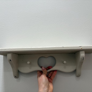 May include: A distressed, off-white wooden shelf with a heart-shaped cutout and two pegs. The shelf is supported by two curved brackets. The shelf is designed for wall mounting and is ideal for displaying small items.