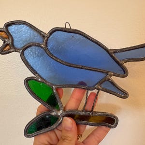 May include: A stained glass suncatcher shaped like a blue bird perched on a branch. The bird features light blue textured glass for the body and head, with a small amber-colored beak. The branch is made of dark green and brown glass pieces.