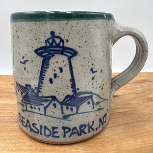 May include: A speckled light gray ceramic mug with a dark green rim and a handle. The mug features a blue hand-painted illustration of a lighthouse and small buildings, with the text "SEASIDE PARK, NJ" written in blue along the bottom edge.