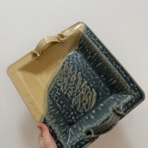 May include: A square ceramic serving dish with a two-tone design. The top half is beige, and the bottom half is a dark blue-green with a textured pattern. It has two curved handles on opposite sides.