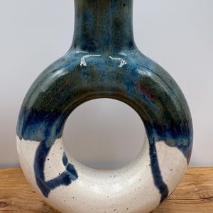 May include: A ceramic vase with a unique circular design. The vase features a dark blue top that transitions to a lighter blue and white base. The vase has a speckled texture and a dark brown interior.