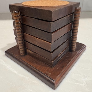 May include: A stack of square dark brown wooden coasters held in a matching wooden holder with vertical ribbed side supports. The top coaster features a circular cork insert. The entire set rests on a square wooden base with beveled edges.