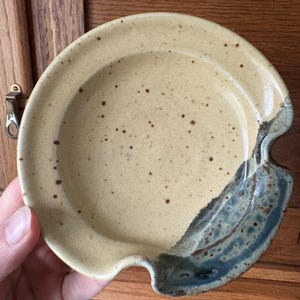 May include: A speckled, ceramic spoon rest with a light tan base and a dark blue and brown accent on the side. The spoon rest has a curved shape with a small notch for the spoon handle.