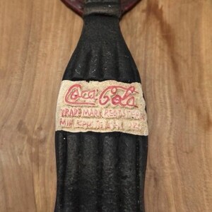 May include: A black cast iron wall-mounted bottle opener shaped like a Coca-Cola bottle. The opener features a circular top plate with the Coca-Cola logo and a central label area displaying the text Coca-Cola Trade Mark Registered.