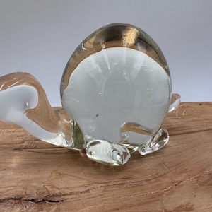 May include: A clear glass turtle figurine with a rounded body and head, and small flipper-like legs. The top of the turtle has a gold-colored rim. The figurine is displayed on a wooden surface.