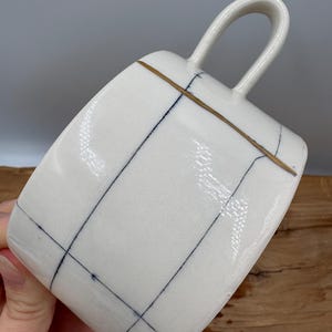 May include: White ceramic mug with a square shape and a curved handle. The mug features a grid pattern of thin, dark blue lines and a gold-colored band near the top. The mug is angled, showcasing its design and form.