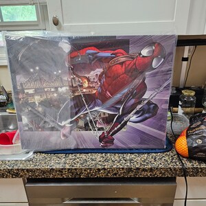 May include: A close-up of a framed print featuring a comic book style illustration of Spider-Man swinging through a city. The print is on a counter top with a bag of oranges in the foreground.