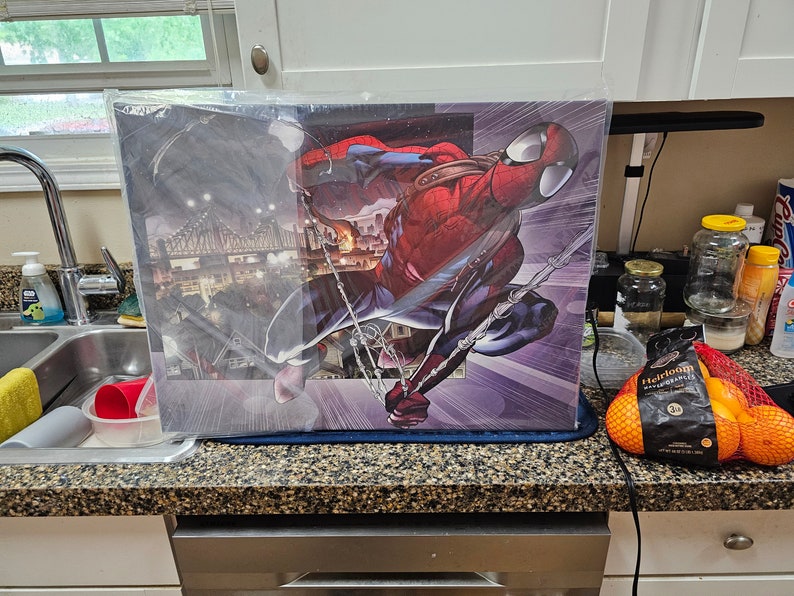 May include: A close-up of a framed print featuring a comic book style illustration of Spider-Man swinging through a city. The print is on a counter top with a bag of oranges in the foreground.