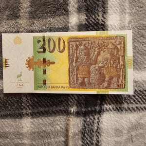 May include: A 200 Macedonian Denar banknote. The bill features a green and yellow design with the number "200" prominently displayed. The right side showcases a detailed relief sculpture. The text "НАРОДНА БАНКА НА РЕПУБЛИКА МАКЕДОНИЈА" is printed on the bill.