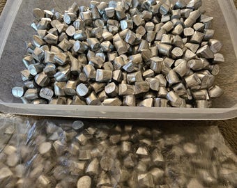 May include: A clear plastic rectangular container filled with numerous small, irregular, silver-colored metal pellets. Below the container, a transparent plastic bag contains a similar quantity of the same silver-colored metal pellets.