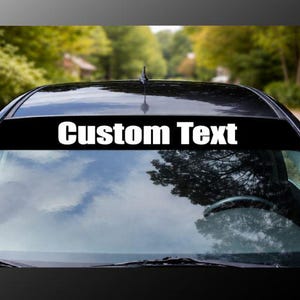 May include: A black car windscreen sun visor banner decal featuring the white text Custom Text centred horizontally. The rectangular adhesive strip is applied to the top portion of the glass, spanning the width of the vehicle windscreen.