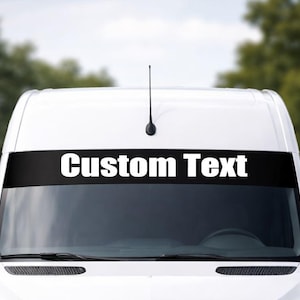Pode incluir: Uma carrinha branca com uma faixa preta no para-brisas. A faixa exibe as palavras "Custom Text" em letras brancas em negrito. A carrinha tem uma antena preta e espelhos laterais.