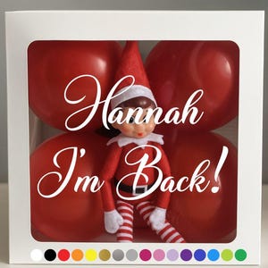 May include: A white box with a clear window showing a red-suited elf doll with the text "Hannah I'm Back!" in white script. The background features large red spheres.