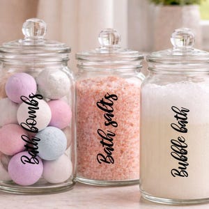 May include: Three glass jars with glass lids, labelled "Bath bombs", "Bath salts", and "Bubble bath". The jar on the left contains pastel-coloured bath bombs. The middle jar holds pink bath salts. The jar on the right is filled with bubbly liquid.
