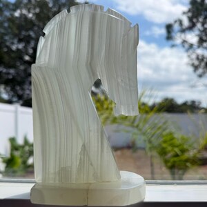 Puede incluir: Una escultura transl&uacute;cida de cabeza de caballo blanquecina con estr&iacute;as verticales. La escultura est&aacute; montada sobre una base rectangular. El fondo muestra una ventana con vistas a un cielo azul y follaje verde.