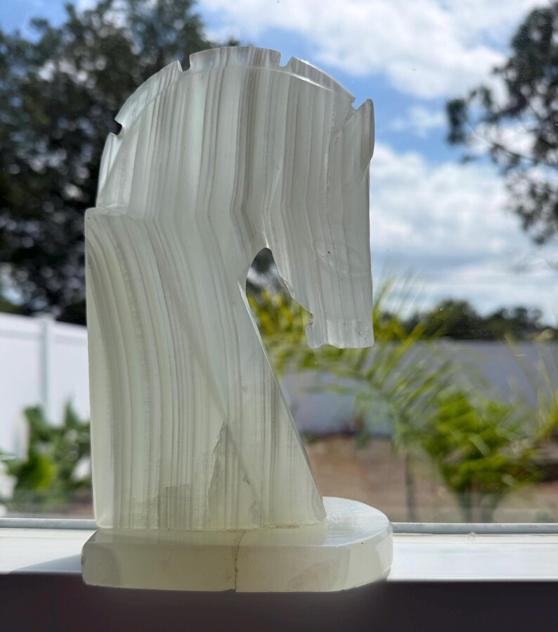 Puede incluir: Una escultura transl&uacute;cida de cabeza de caballo blanquecina con estr&iacute;as verticales. La escultura est&aacute; montada sobre una base rectangular. El fondo muestra una ventana con vistas a un cielo azul y follaje verde.