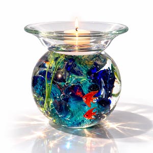 Może przedstawiać: Przezroczysty szklany świecznik z okrągłą podstawą wypełnioną niebieskimi i zielonymi kamieniami dekoracyjnymi oraz dwiema czerwonymi rybami. Mała zapalona świeca znajduje się w przezroczystej szklanej misce na górze.