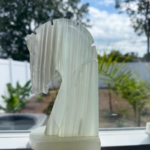 Puede incluir: Escultura de cabeza de caballo transl&uacute;cida blanquecina. La cabeza del caballo est&aacute; tallada con l&iacute;neas verticales y se asienta sobre una base rectangular. La escultura est&aacute; colocada en un alf&eacute;izar con vistas a &aacute;rboles y cielo azul.