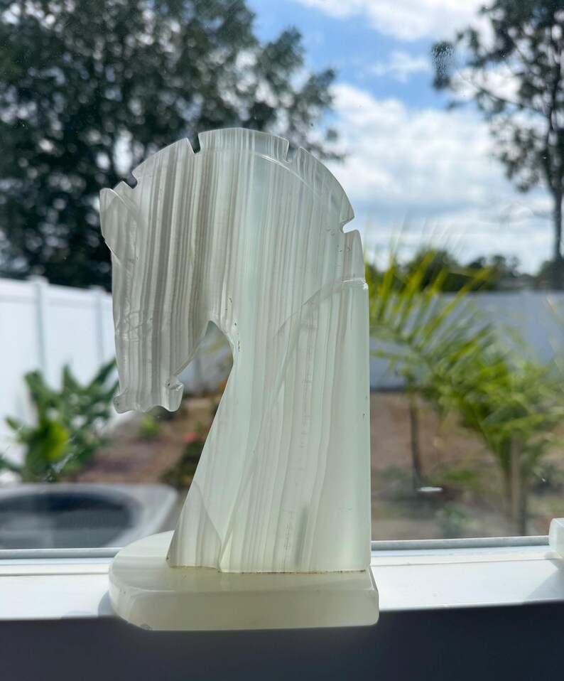 Puede incluir: Escultura de cabeza de caballo transl&uacute;cida blanquecina. La cabeza del caballo est&aacute; tallada con l&iacute;neas verticales y se asienta sobre una base rectangular. La escultura est&aacute; colocada en un alf&eacute;izar con vistas a &aacute;rboles y cielo azul.