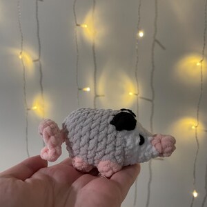 Puede incluir: Un pequeño juguete de cerdo de crochet hecho con hilo rosa, blanco y gris. El cerdo tiene la nariz y los ojos negros.