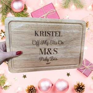 Puede incluir: Una tabla de cortar rectangular de madera con un borde empotrado. La tabla tiene el texto grabado "KRISTEL Off My Tits On M&S Picky Bits". La tabla está sostenida por una mano con esmalte de uñas rojo. Adornos y regalos rosas en el fondo.